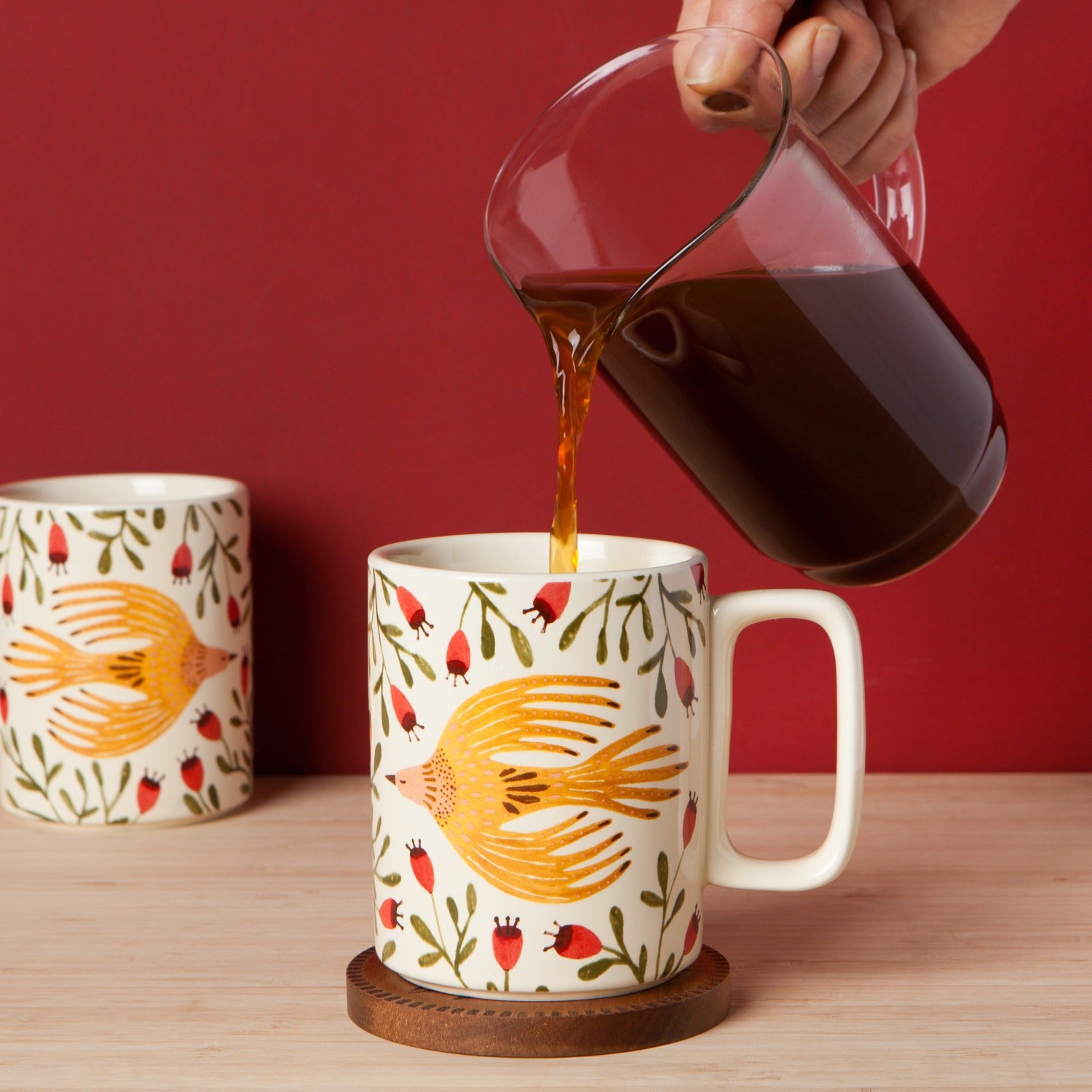 Person pouring coffee into a mug with bird design on a wooden table against a red background
