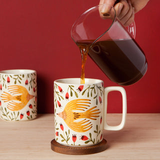 Person pouring coffee into a mug with bird design on a wooden table against a red background