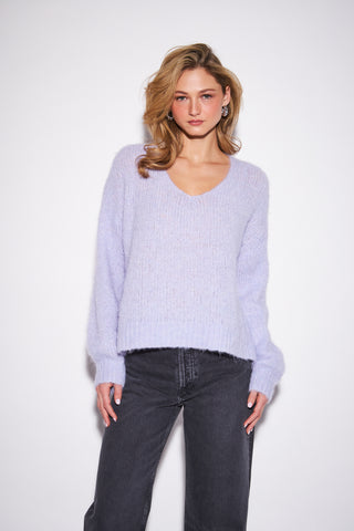 A woman modeling a light lavender fuzzy knit V-neck sweater with dark gray wide-leg jeans, standing against a plain white studio background.