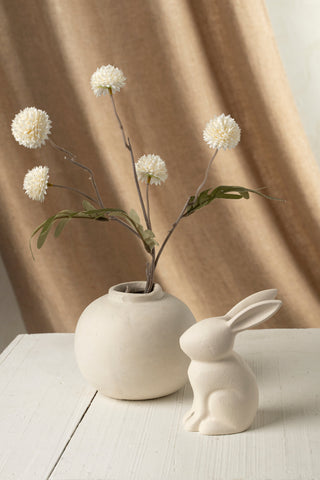 Small white vase with flowers and a ceramic rabbit figurine on a light surface with a beige curtain background.
