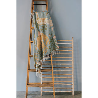 Decorative blanket with fringe draped over a wooden ladder against a gray wall.