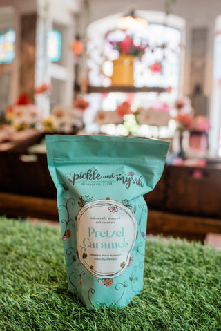 Teal package of 'Pickle and Myrsh' Pretzel Caramels on a grass-like surface with a blurred indoor background.