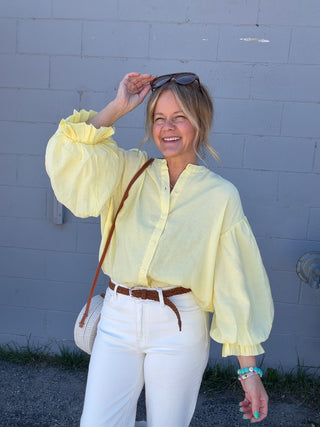 Woman wearing a yellow blouse and white pants against a gray wall.