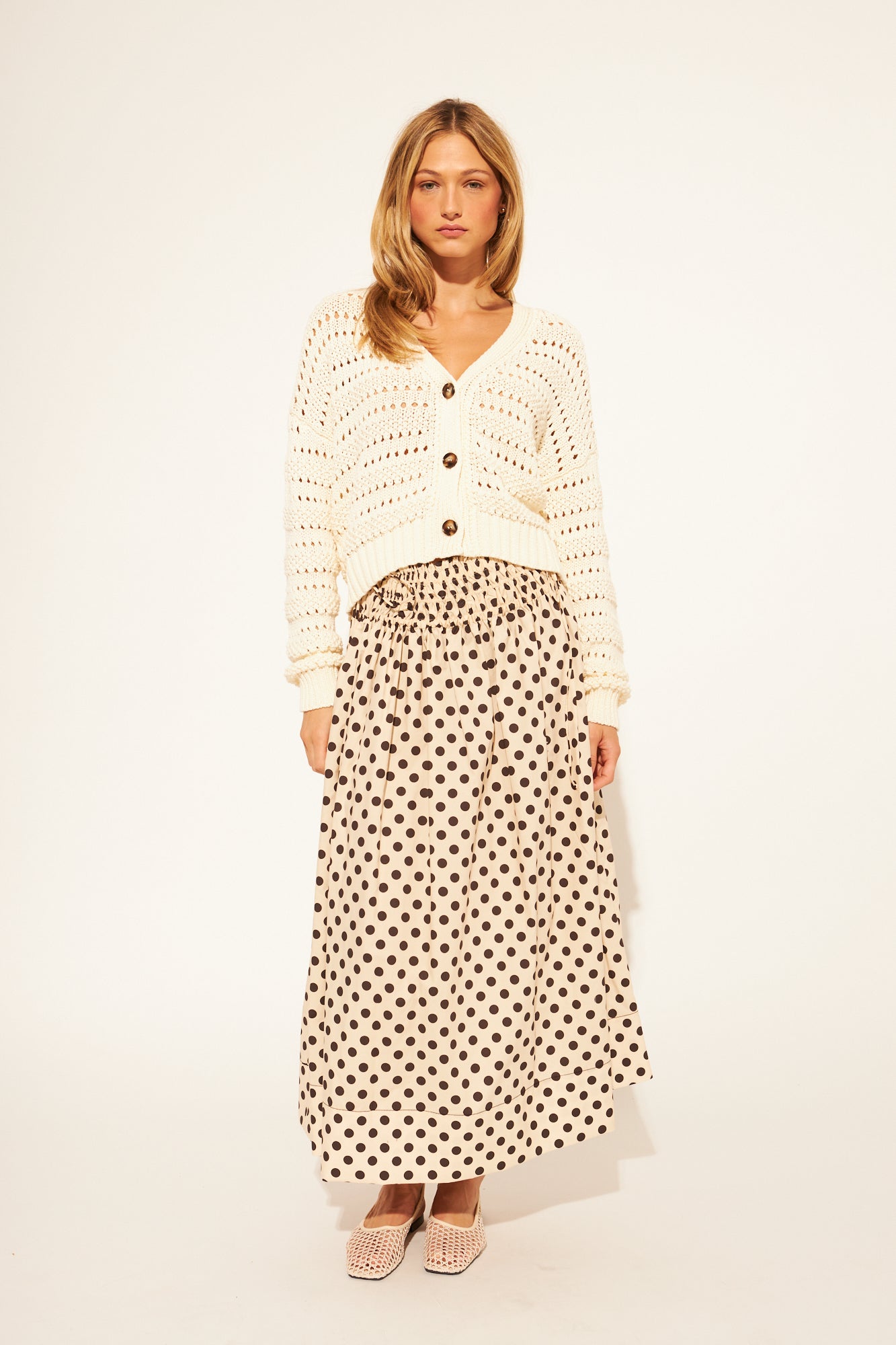 Woman wearing a white knit cardigan and polka dot skirt on a plain background
