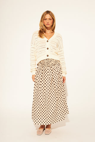 Woman wearing a white knit cardigan and polka dot skirt on a plain background