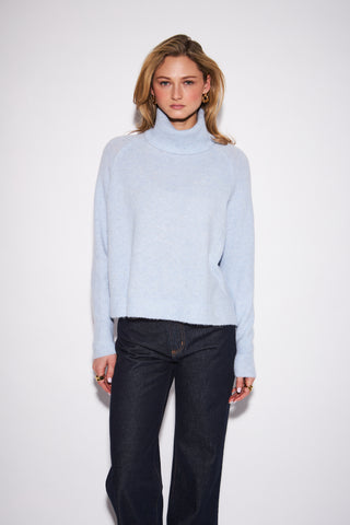 A woman modeling a light blue turtleneck sweater and dark blue jeans against a plain white studio background.