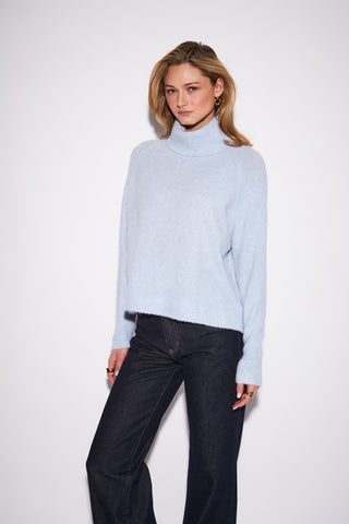 A woman modeling a light blue turtleneck sweater with dark blue jeans against a plain white studio background.