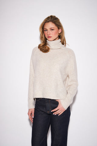 A woman modeling a light beige knit turtleneck sweater paired with dark wash jeans, standing against a plain white studio background.
