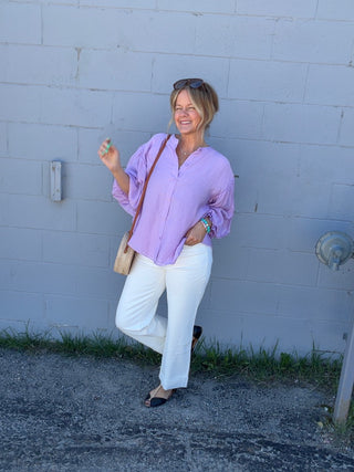 Woman in a purple shirt and white pants standing against a light blue wall.