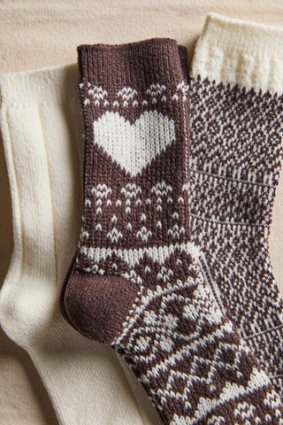 Three knitted socks arranged on a beige fabric background: one cream sock, one dark brown sock with a large white heart and white Fair Isle pattern, and one dark brown and cream patterned sock.