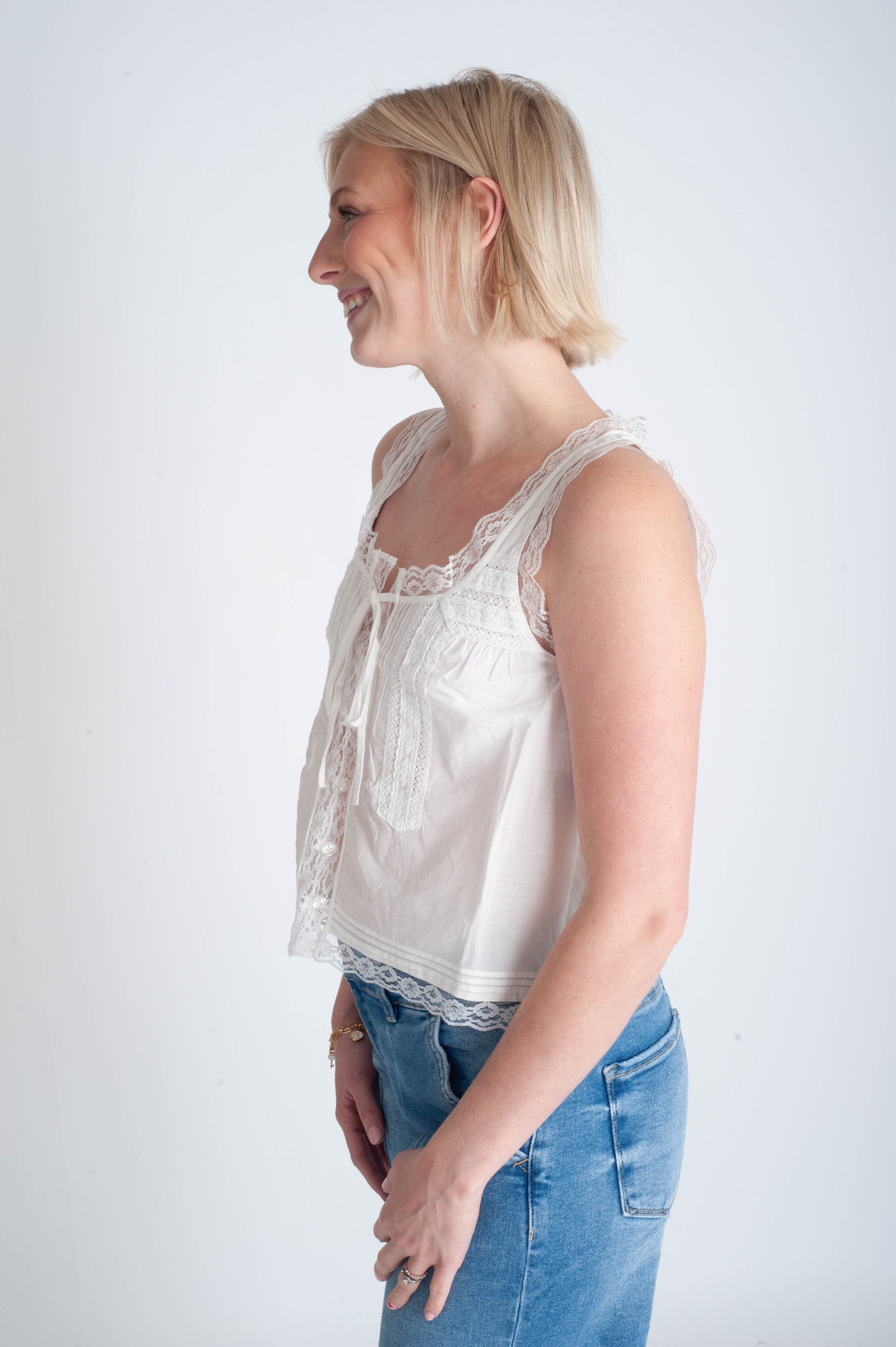 Woman wearing a white sleeveless top with lace details and blue jeans on a white background