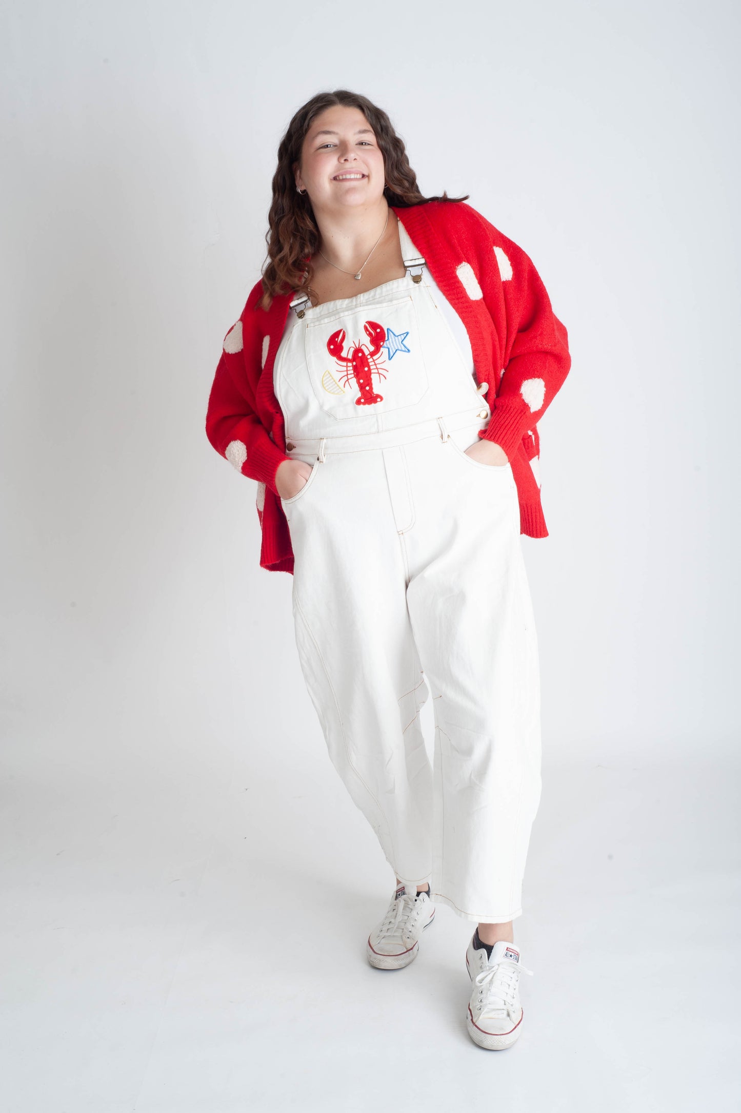 Person wearing a red cardigan, white overalls with a lobster design, and white sneakers on a white background