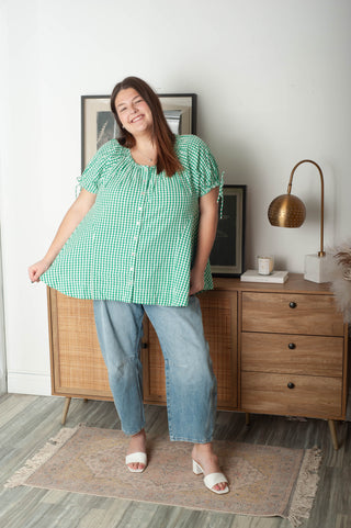 Woman in a green checkered shirt and blue jeans standing in a room with wooden furniture and decor.