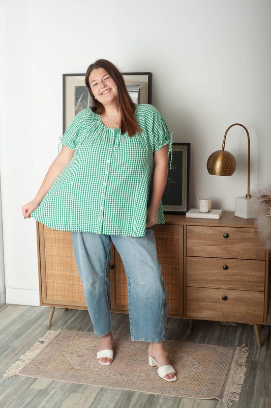 Woman in a green checkered shirt and blue jeans standing in a room with wooden furniture and decor.