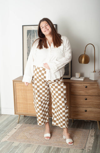 Woman in a white blouse and checkered pants standing in a room with wooden furniture and decor.