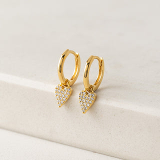 Gold hoop earrings with heart-shaped diamond pendants on a beige surface