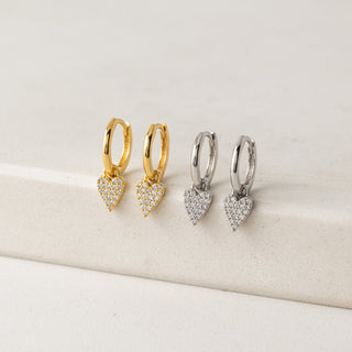 Two pairs of heart-shaped hoop earrings in gold and silver on a beige surface.
