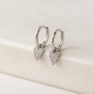 Silver hoop earrings with heart-shaped diamond pendants on a beige surface