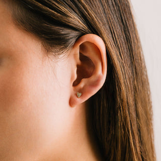 A person wearing a pair of gold-plated stud earrings with cubic zirconia stones, displayed on a white surface.