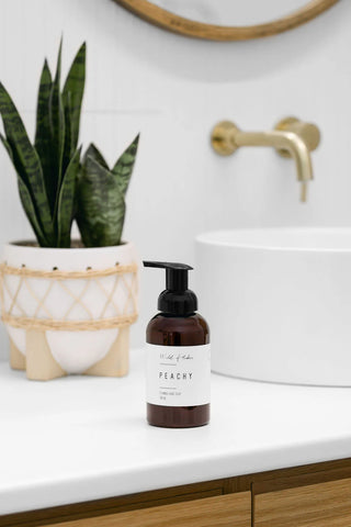 Bottle of 'Peach' soap on a bathroom counter with a plant and sink in the background.