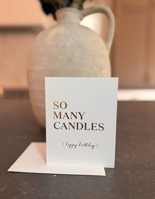 Card with 'So Many Candles (Happy Birthday)' text in front of a ceramic vase on a table.