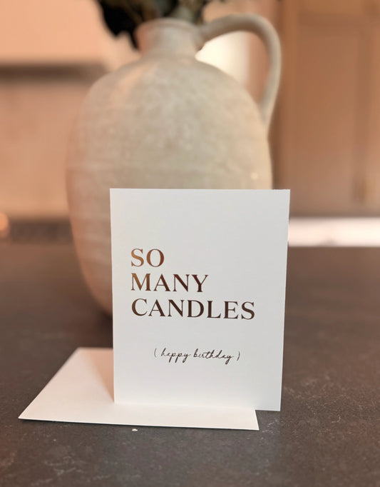 Card with 'So Many Candles (Happy Birthday)' text in front of a ceramic vase on a table.