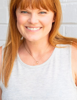 Woman with red hair wearing a gray tank top against a white brick wall.