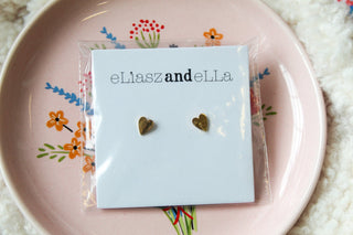 A pair of gold-colored heart-shaped earrings on a white backing, presented on a pink plate with floral patterns.