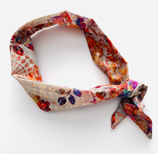 A lightweight fabric neck scarf with a multicolor abstract floral and geometric print on a cream base, styled in a loop with a knot at the end, photographed on a clean white background.