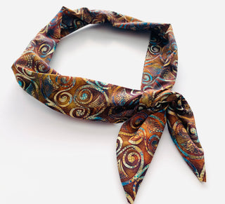 A patterned silk scarf with a brown, gold, and blue paisley swirl design, tied in a knot on a plain white background.