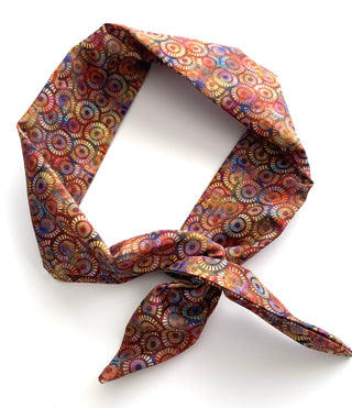 A patterned neck scarf tied in a knot, featuring a dense circular mandala print in shades of red, orange, yellow, blue, and brown on a white background.