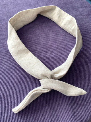 A light beige linen neck gaiter or headband tied in a knot, displayed on a textured purple background.