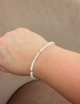 A delicate beaded bracelet with small white pearls and tiny gold spacer beads, shown worn on a person's wrist against a neutral brown background.