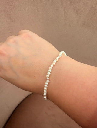 A delicate white beaded bracelet worn on a person's wrist, shown against a neutral brown background.