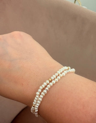 A close-up of a person's wrist wearing two strands of white freshwater pearl bracelets with small gold spacer beads, set against a soft beige background.