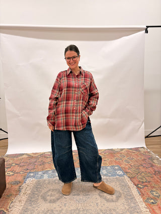A woman wearing a red and brown plaid button-down shirt, wide-leg dark blue jeans, and tan slip-on shoes stands in front of a white studio backdrop on a patterned rug.