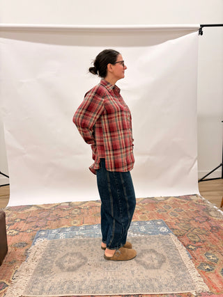 A woman stands in profile wearing a red and green plaid button-up shirt, dark blue jeans, and tan slip-on shoes, standing on a patterned rug in front of a white studio backdrop.