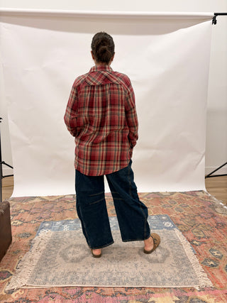 A person standing with their back to the camera, wearing a red and dark plaid flannel shirt and wide-leg dark blue jeans, standing on a patterned rug in front of a white studio backdrop.