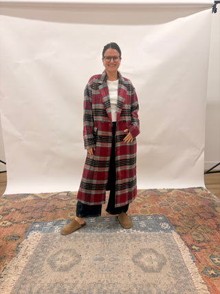 A woman wearing a long red and black plaid coat over a white top and black pants, standing in front of a white studio backdrop on a patterned rug.