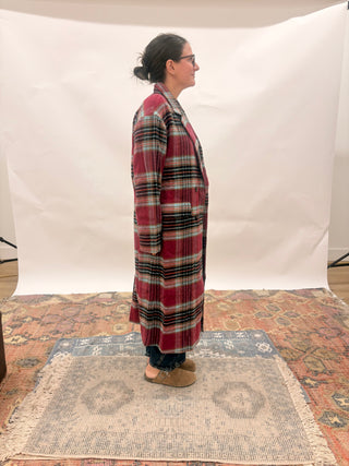 A person stands in profile wearing a long red plaid coat over dark pants and tan shoes, posed on a patterned rug in front of a white studio backdrop.