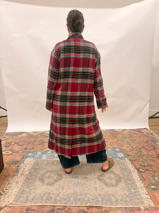 A person viewed from behind wearing a long red plaid coat over dark pants, standing on a patterned rug in front of a white studio backdrop.