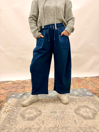 A person wearing dark navy wide-leg drawstring pants, a gray knit sweater, and light-colored sneakers, standing on a patterned rug in front of a neutral backdrop.