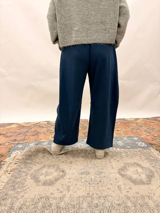 A person shown from the back wearing dark navy wide-leg trousers, a gray knit sweater, and light-colored shoes, standing on a patterned rug against a plain off-white backdrop.