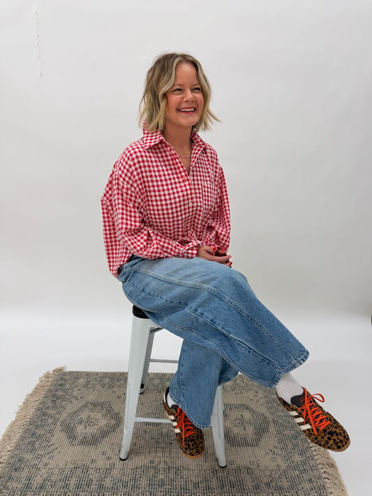 Person wearing a red checkered shirt and blue jeans sitting on a stool against a white wall.