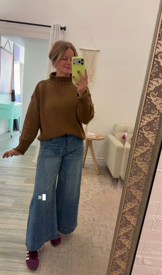 A woman takes a mirror selfie in a room, wearing a brown mock-neck knit sweater and wide-leg blue jeans.
