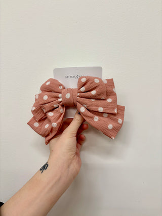 A hand holding a black hair bow with white polka dots, with a visible brand tag on the top left corner.