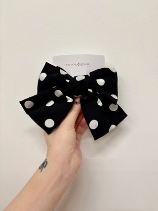 A hand holding a black hair bow with white polka dots, with a visible brand tag on the top left corner.