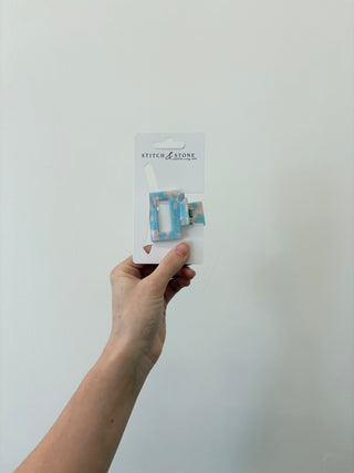 A hand holding a square-shaped green and beige hair clip, packaged in a white plastic container with the brand 'Stitch & Stone' visible on it.
