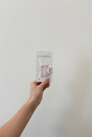 A hand holding a square-shaped green and beige hair clip, packaged in a white plastic container with the brand 'Stitch & Stone' visible on it.