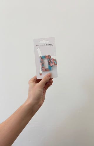 A hand holding a square-shaped green and beige hair clip, packaged in a white plastic container with the brand 'Stitch & Stone' visible on it.